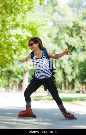 Una donna gioiosa che gira a rotelle in un parco, bilanciata e sorridente, con alberi che forniscono uno sfondo verde lussureggiante. Indossa abiti estivi casual e porta con sé Foto Stock