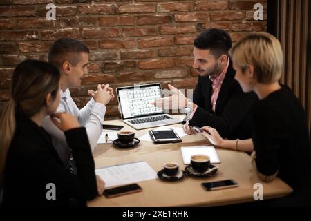 Quattro professionisti si sono riuniti intorno a un notebook in un bar con pareti a mattoni, discutendo delle strategie aziendali anziché del caffè. Atmosfera coinvolgente e mirata. Foto Stock