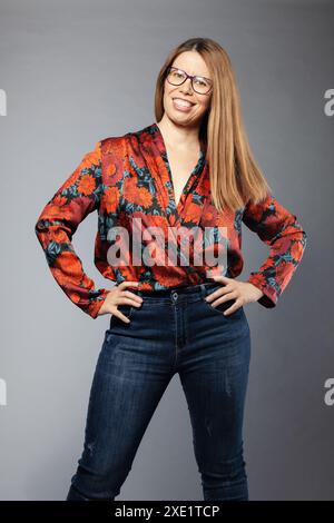 Una giovane donna vivace ed elegante che indossa una blusa floreale e un paio di jeans è in piedi con le mani sui fianchi su uno sfondo grigio. Foto Stock