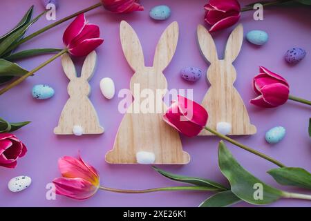 Uova di Pasqua e coniglietti di legno con tulipani rosa su sfondo viola pastello. Composizione piatta, vista dall'alto. Decorazioni minimali Foto Stock