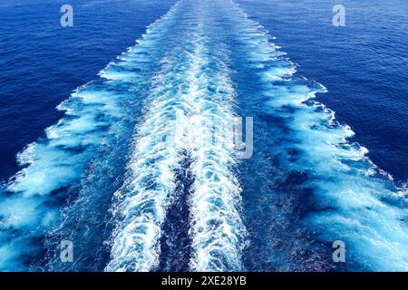 Wide turbulent white foam water trail behind a large cruise ship in a calm blue sea on a sunny day Foto Stock