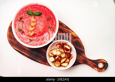 Zuppa fredda di pomodoro vegetale con basilico e sedano su una tavola di legno su uno sfondo chiaro con crostini in una ciotola bianca. Foto di alta qualità Foto Stock