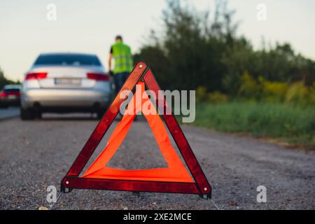 Incidente d'emergenza auto di lusso in argento rotto. Uomo conducente che installa un segnale di stop triangolare rosso su strada. Auto sportiva accesa Foto Stock