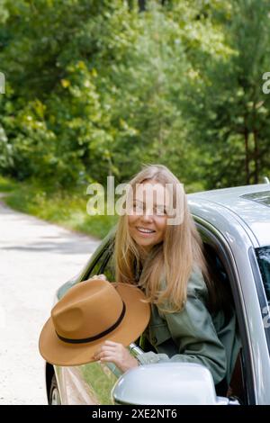 Donna bionda con cappello che attacca la testa fuori dall'auto con parabrezza. I giovani turisti esplorano i viaggi locali rendendo i momenti reali. Vero emo Foto Stock