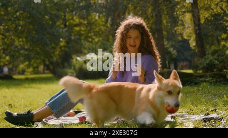 Ragazza caucasica che gioca con il cane simpatico cucciolo giocoso in città all'aperto gestore donna che dà da mangiare all'animale domestico corgi gallese dorati con picnic estivo di frutta Foto Stock