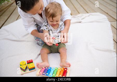 Una madre felice e il suo bambino giocano con giocattoli musicali colorati su una coperta all'aperto, favorendo la creatività e l'adesione. Foto Stock