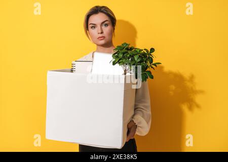 Donna che tiene una scatola con oggetti personali dopo le dimissioni dal lavoro su sfondo giallo Foto Stock