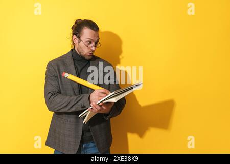 Uomo divertente che indossa occhiali con una matita grande e un taccuino su sfondo giallo Foto Stock