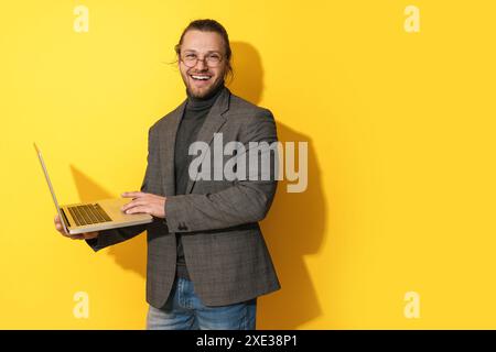 Un uomo con la barba felice che indossa gli occhiali da vista sta utilizzando un computer portatile su sfondo giallo Foto Stock