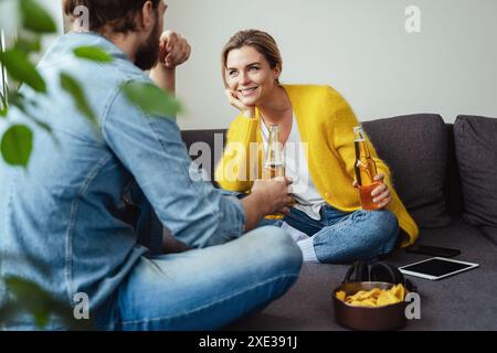 Una giovane e allegra coppia seduta sul divano bevendo birra e mangiando nachos Foto Stock