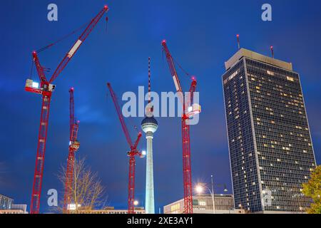 La famosa torre della televisione di Berlino di notte con un grattacielo e quattro gru rosse Foto Stock