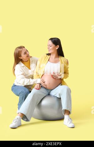 Doula matura con donna incinta su fitball su sfondo giallo Foto Stock