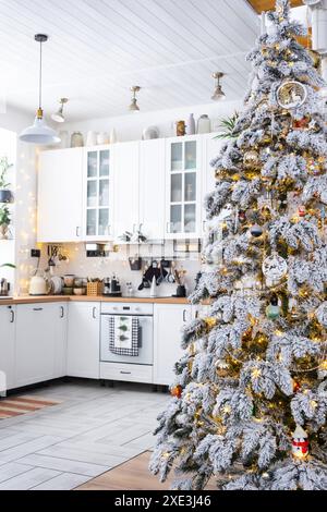 Decorazioni natalizie natalizie in cucina bianca, interni moderni in stile rustico con un albero di Natale innevato e luci fiabesche. Anno nuovo, Christm Foto Stock