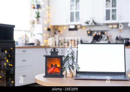 Computer portatile con schermo bianco ripreso nell'accogliente cucina di Natale decorata di colore bianco con luci delle fate e un albero di Natale. Seasona Foto Stock