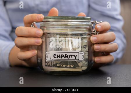 Mani femminili che risparmiano denaro in vasetti di vetro pieni di banconote in dollari. Trascrizione DI BENEFICENZA davanti al barattolo. Gestione f. Personale Foto Stock