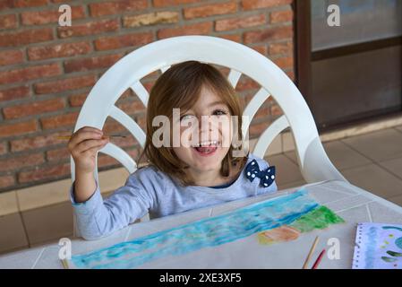 ragazza che dipinge un disegno ad acquerello tenendo in mano un pennello e sorridente, con un muro di mattoni sullo sfondo Foto Stock