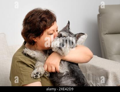 Ritratto di una donna anziana che bacia un cane schnauzer in miniatura sul divano Foto Stock