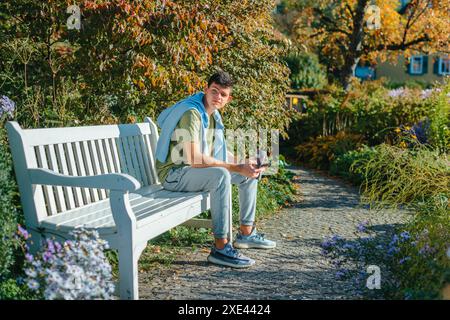 Un adolescente siede su Una panchina nell'Autumn Park beve un caffè da Una tazza Thermo e guarda Un telefono. Ritratto di Handsome Cheer Foto Stock