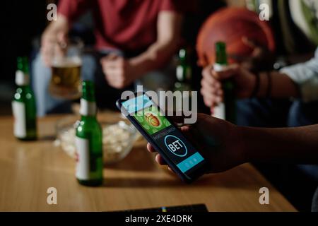Uomo irriconoscibile che tiene in mano lo smartphone scommettere sulla squadra preferita mentre guarda la partita di basket con i suoi amici la sera Foto Stock