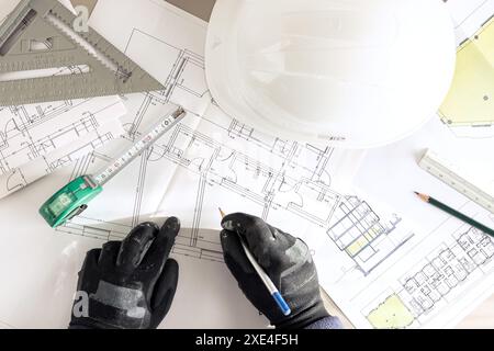 Vista dall'alto di un appaltatore che lavora con una matita sui piani di architettura sulla scrivania di un cantiere edile Foto Stock