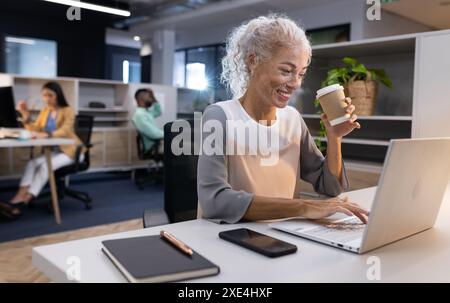 Lavora in ufficio, una donna anziana che usa un computer portatile e beve un caffè alla scrivania Foto Stock