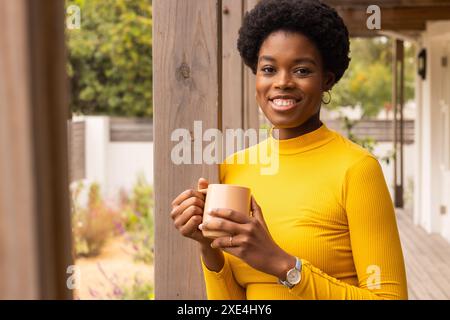 Donna sorridente in maglione giallo che tiene una tazza di caffè sul portico, godendosi la mattina Foto Stock