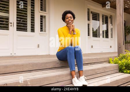 Seduta sui gradini della veranda, donna in maglione giallo sorridente e rilassante all'aperto Foto Stock
