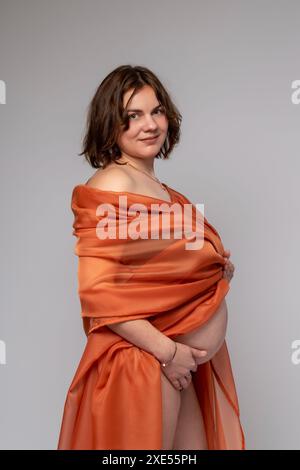 Una donna incinta in un mantello arancione tiene le mani sullo stomaco su uno sfondo grigio chiaro. Gravidanza, maternità, preparazione e aspettativa di un Foto Stock