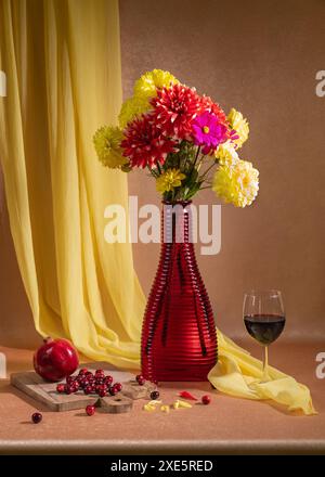 Ci sono molti fiori luminosi in un vaso rosso, un bicchiere di vino rosso si trova nelle vicinanze, bacche rosse e melograni giacciono. Sfondo arancione e handker giallo Foto Stock