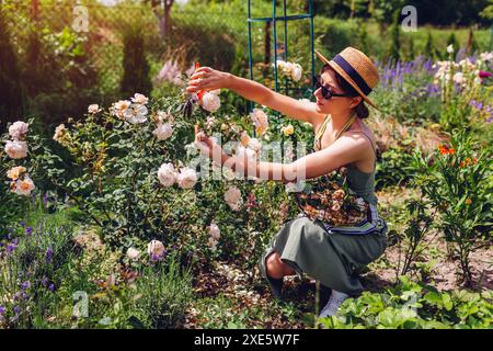La giovane donna che ha trascorso la morte ha trascorso la fioritura delle rose nel giardino estivo. Il giardiniere taglia i fiori secchi con la potatrice e li mette nel cestino. Cottage inglese g Foto Stock