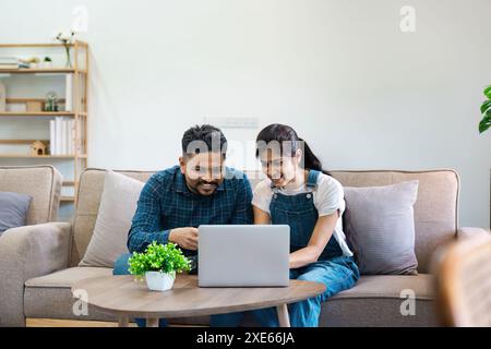 Giovane coppia indiana che lavora insieme sul laptop a casa, sorride e discute di idee in un salotto moderno Foto Stock