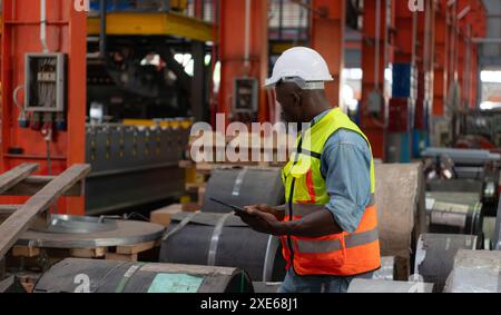 Vista frontale di un giovane lavoratore che utilizza un tablet digitale per controllare le scorte di lamiere in fabbrica Foto Stock