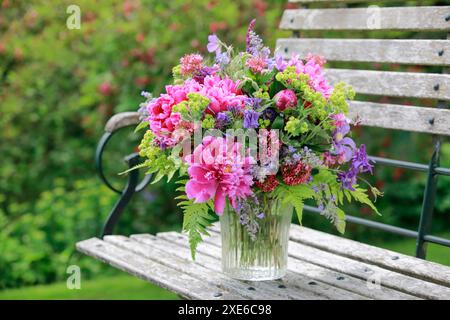 Bouquet colorato di fiori rossi, rosa e viola con peonie e columbine in un vaso di vetro su una panchina da giardino in legno Foto Stock