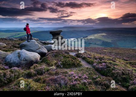 Walker si affaccia dalla caratteristica formazione rocciosa di Salt Cellar su una brughiera di heather in estate, Derwent Edge, Peak District National Park, Derbysh Foto Stock