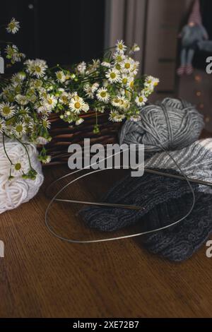 Palle in filato e calze lavorate a maglia con cesto di margherite bianche su tavolo di legno. Hobby per lavorare a maglia. Lana grigia e bianca e aghi per maglieria con calzini caldi. Foto Stock