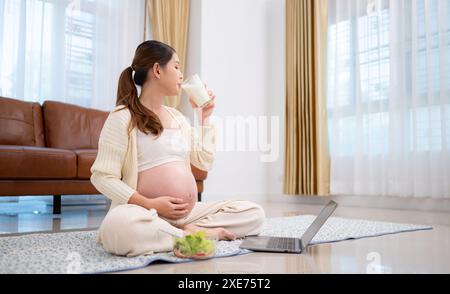 Donna incinta che beve latte seduto sul pavimento a casa Foto Stock