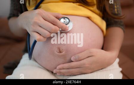 Donna asiatica incinta che ascolta il battito cardiaco del bambino con stetoscopio a casa Foto Stock