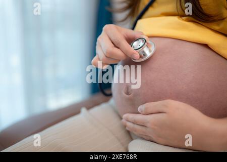 Donna asiatica incinta che ascolta il battito cardiaco del bambino con stetoscopio a casa Foto Stock