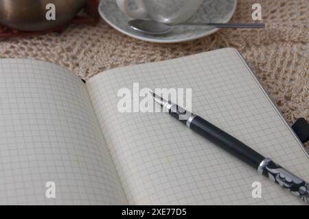 Una penna su un foglio bianco del suo diario aperto accanto a una tazza di caffè su un tavolo con un piatto all'uncinetto Foto Stock