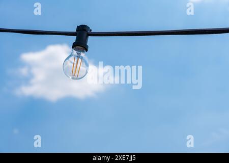 Un primo piano di una singola lampadina appesa su un filo contro un cielo azzurro limpido in una giornata di sole, catturando l'essenza della semplicità e del design moderno Foto Stock