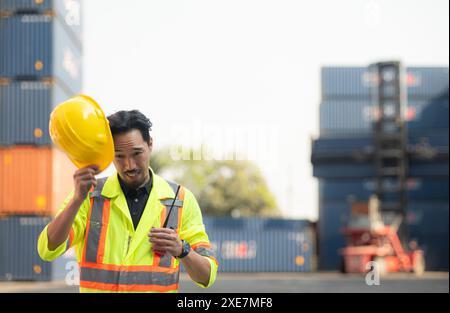 Ritratto di un lavoratore asiatico maschile che indossa un giubbotto di sicurezza e un elmetto, che si prende una pausa dal lavoro con una scatola portacontainer nel retro Foto Stock