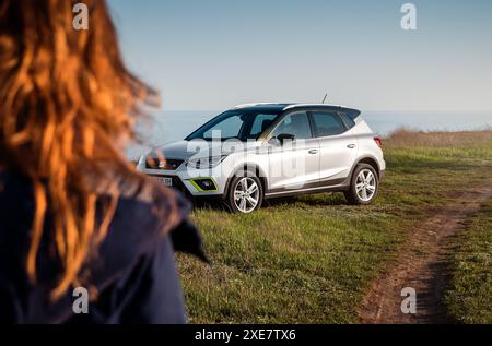 Donna che si avvicina a un SEDILE argentato Arona parcheggiata su un'erba su un lato della strada sterrata - oltre la vista delle spalle. Vista frontale di tre quarti della berlina spagnola. Foto Stock