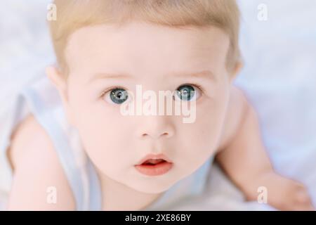 Il bambino con gli occhi blu giace su una coperta bianca, guardando in alto curiosamente, irradiando innocenza e purezza. Questo coinvolgente ritratto cattura la gioia di Foto Stock