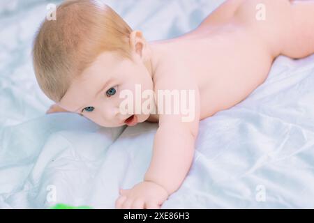 Un bambino felice gattona su un comodo letto con lenzuola bianche, guardando curiosamente con grandi occhi blu, circondato dall'amore in una camera da letto luminosa Foto Stock