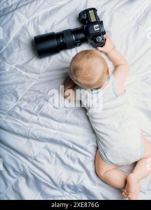 Il bambino giace su una trapunta bianca, raggiungendo una fotocamera di grandi dimensioni. La scena suggerisce umoristicamente un futuro in cui potrebbe dire: "Voglio essere disoccupato quando ho un Foto Stock