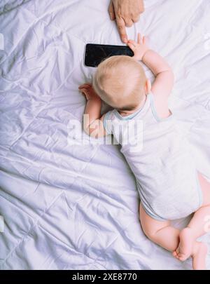 Il bambino giace su una trapunta bianca, raggiungendo uno smartphone guidato dalla mano di un adulto. Questa scena incarna il concetto di istruzione come guida attraverso la nuova c Foto Stock