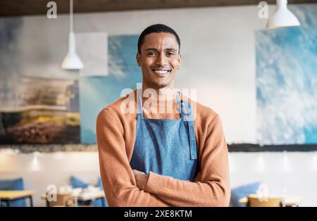 Un giovane marocchino sorridente che indossa un grembiule casual in una caffetteria moderna. E' in piedi con le braccia incrociate. Lo sfondo mostra un ambiente accogliente ed elegante Foto Stock