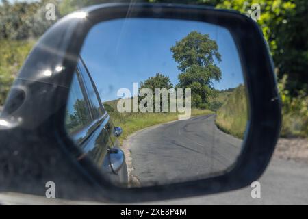 Vista sullo specchietto retrovisore di un'auto che guarda indietro lungo una strada di campagna nel Sussex rurale, in una soleggiata giornata estiva Foto Stock