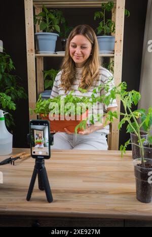 La blogger donna registra video sul suo smartphone nel suo giardino interno, tenendo un vaso di erbe fresche circondato da piante in vaso e attrezzi da giardino. La ragazza sta trasmettendo online come prendersi cura delle piantine. Foto Stock