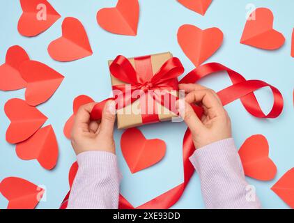 La mano di una donna regge una confezione regalo avvolta da un nastro di seta rosso su sfondo blu Foto Stock